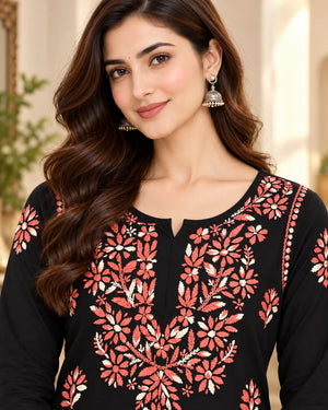 A woman wearing a black long-sleeve kurta with coral pink and cream floral embroidery, paired with gold-toned jhumka earrings, in a softly lit indoor setting.