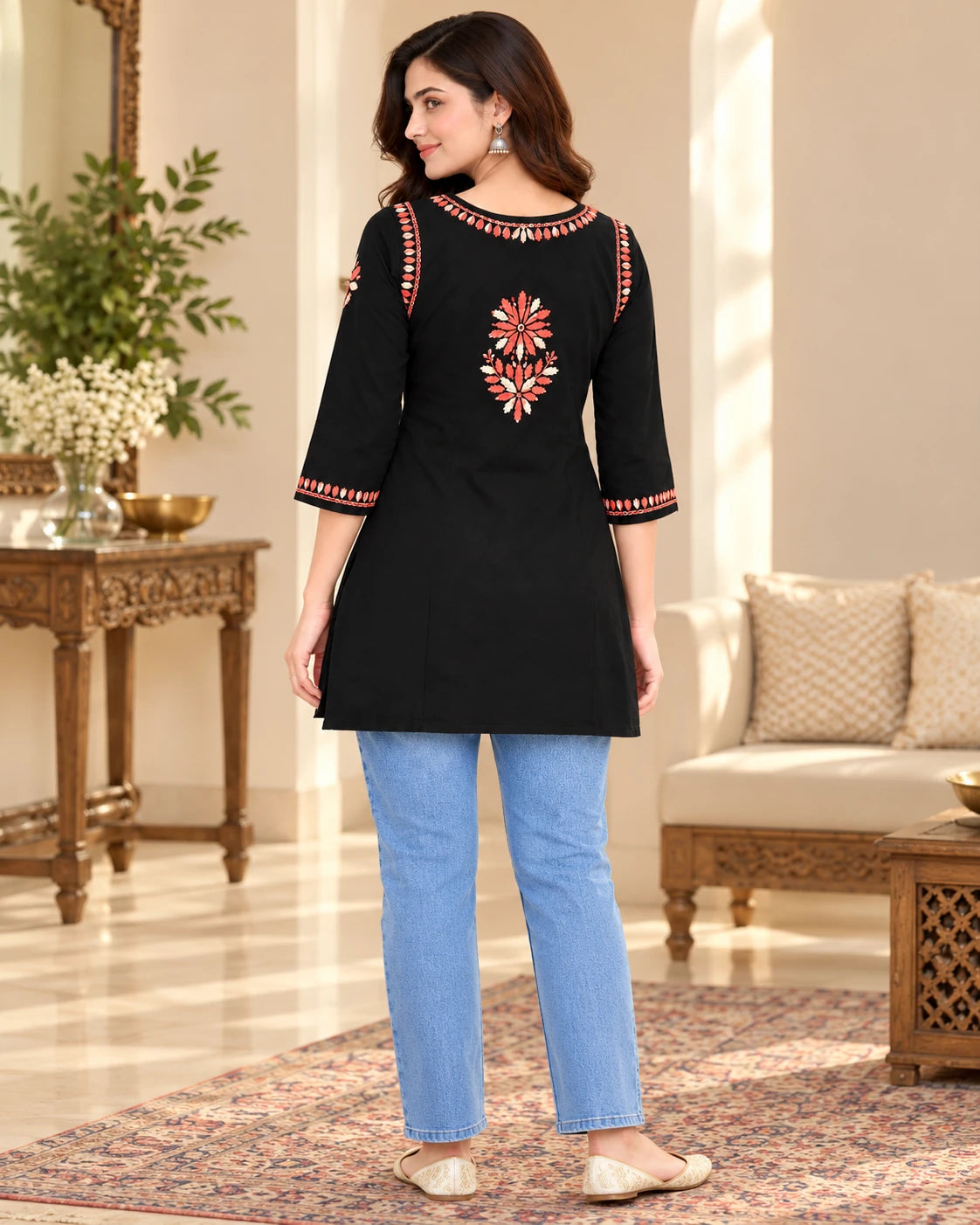 A woman wearing a black three-quarter sleeve kurta with red and gold floral embroidery on the back and sleeve cuffs, paired with light blue jeans, standing in a decorated indoor room.