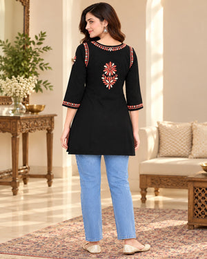A woman wearing a black three-quarter sleeve kurta with red and gold floral embroidery on the back and sleeve cuffs, paired with light blue jeans, standing in a decorated indoor room.