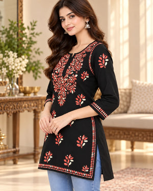 A woman modeling a black three-quarter sleeve kurta with red and white floral embroidery, paired with light blue jeans in a warm, elegant indoor setting.