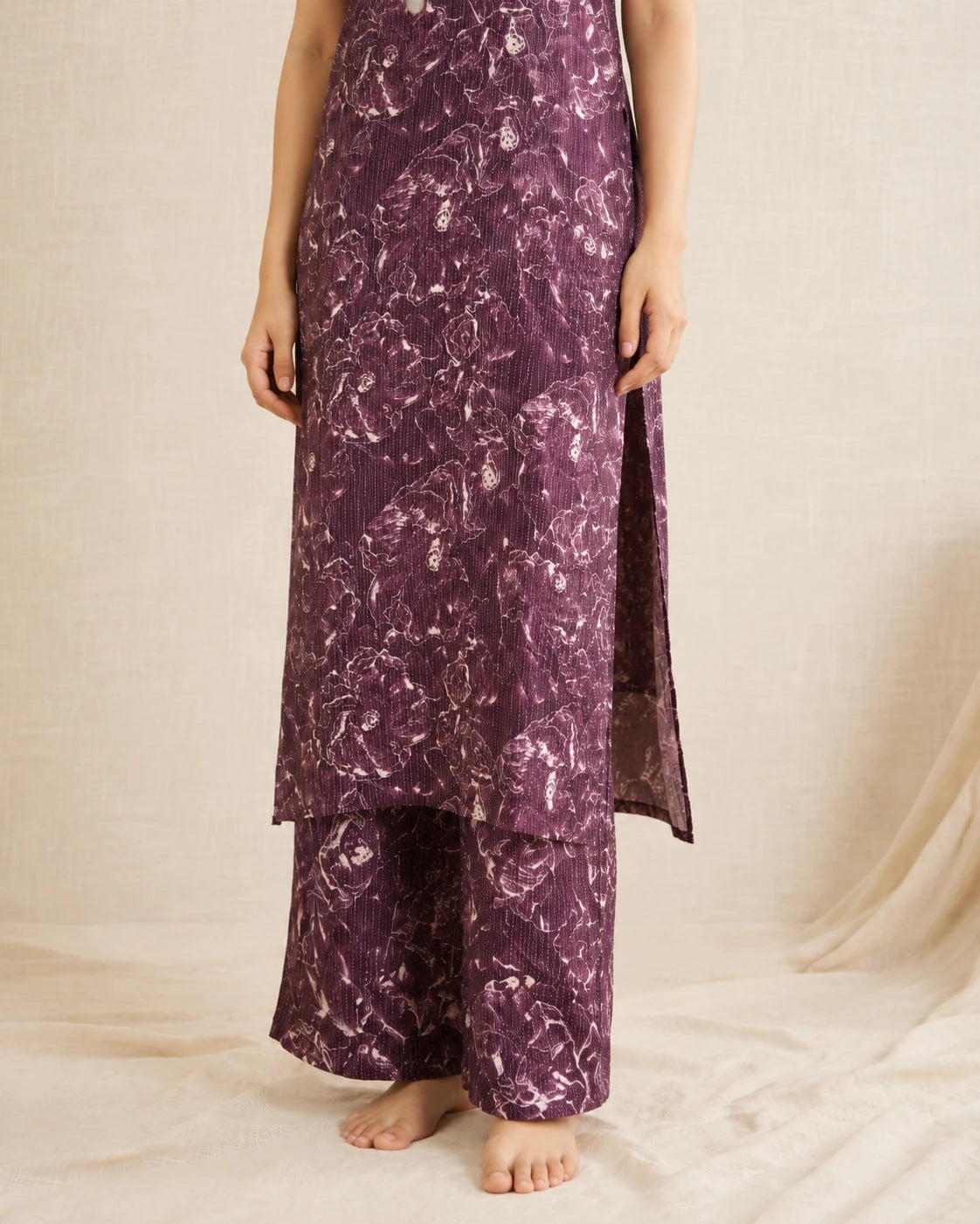 A person wearing a long purple floral printed kurta with matching wide-leg pants, standing barefoot against a draped cream fabric backdrop.