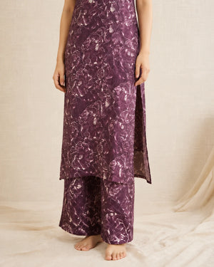 A person wearing a long purple floral printed kurta with matching wide-leg pants, standing barefoot against a draped cream fabric backdrop.