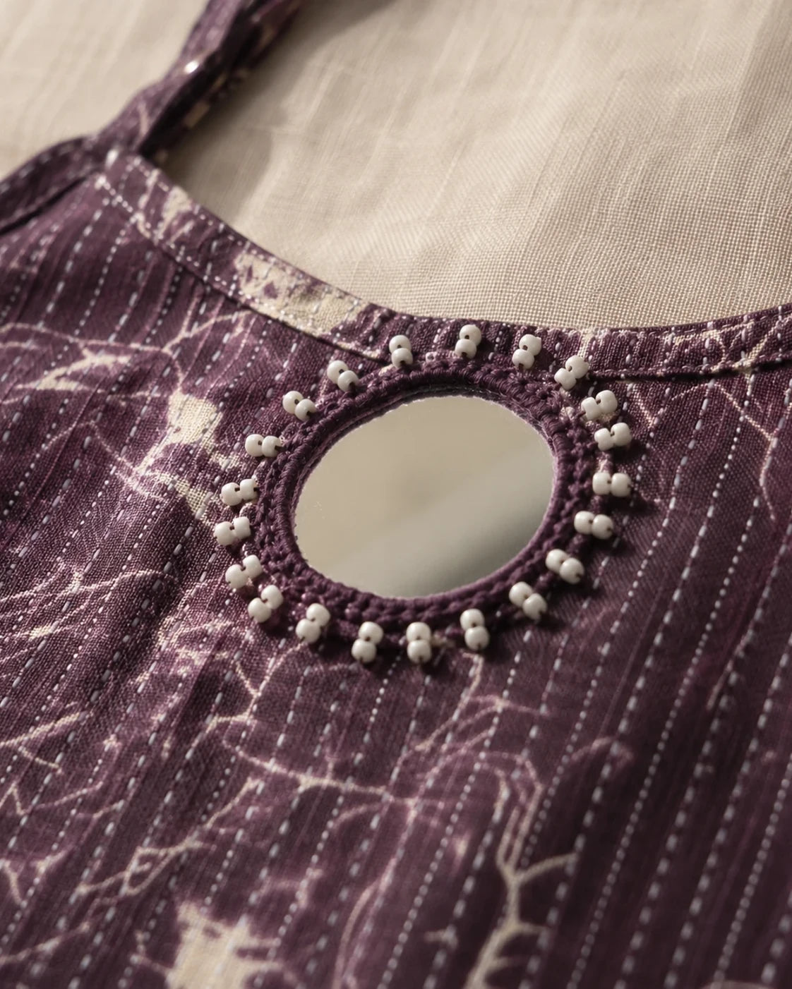 Close-up of a purple patterned garment with a round cutout neckline decorated with a ring of small white beads, set against a light beige fabric background.