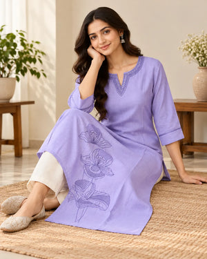A woman sits on a woven jute mat wearing a lavender purple kurta with a split neckline, 3/4 sleeves, and a floral print on the lower front, paired with off-white pants and beige flat shoes in a warm, sunlit indoor setting.