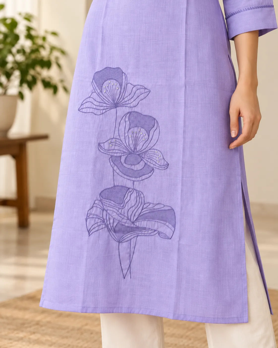 A close-up of a person wearing a lavender purple kurta with a floral embroidery design on the front, paired with off-white trousers in a softly lit indoor setting.