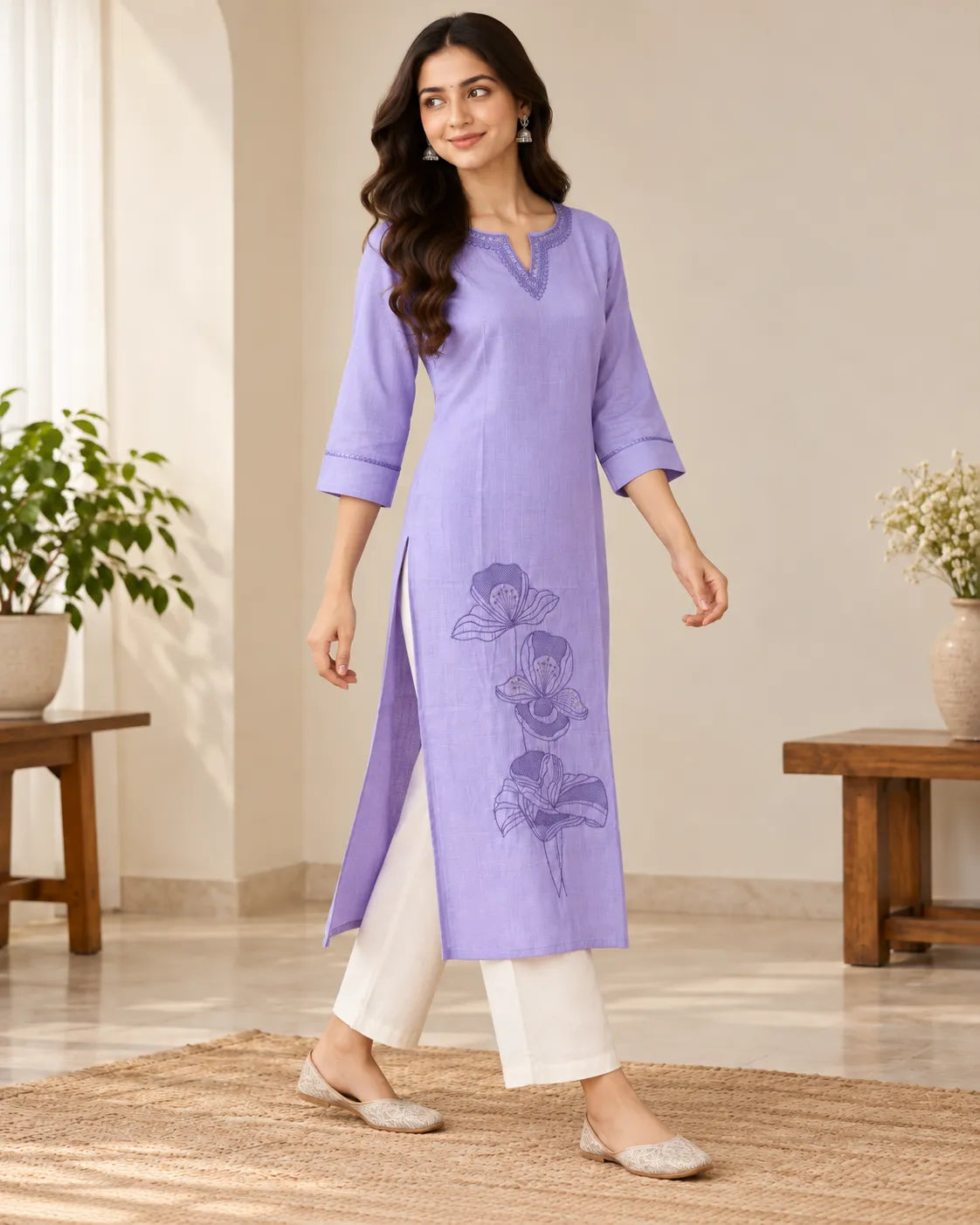 A woman models a lavender purple cotton kurta with a split side slit and floral embroidery, paired with white pants and beige flat shoes in a bright indoor setting.