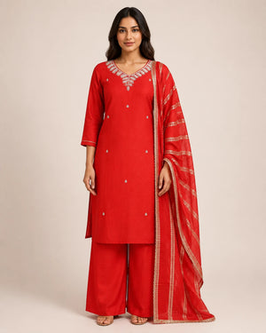 A woman modeling a red Indian ethnic suit set featuring a kurta with silver embroidery, matching wide-leg trousers, and a sheer red dupatta with gold zari borders, set against a plain beige studio background.