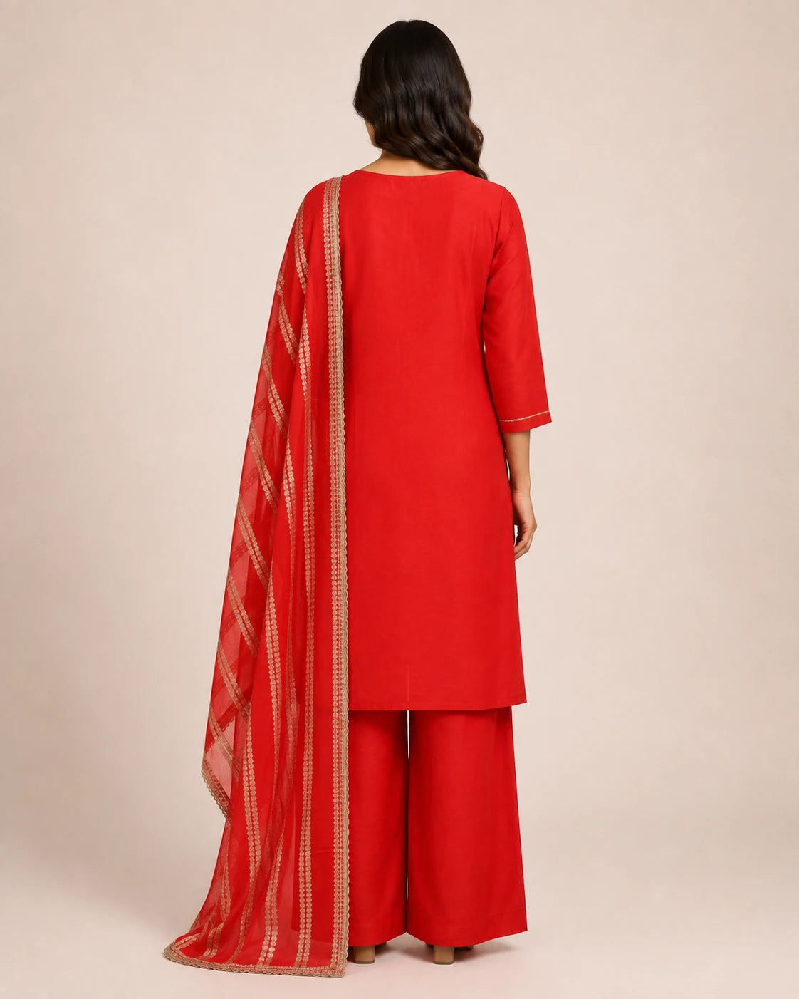 A woman shown from the back wearing a bright red three-piece traditional outfit with matching red trousers and a sheer red dupatta featuring a gold patterned border, set against a plain beige studio background.