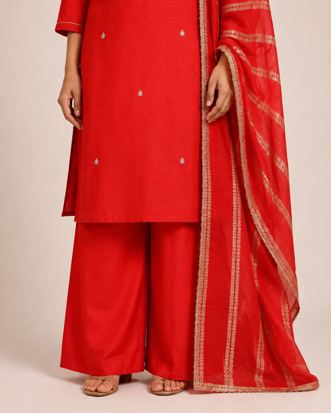 A red traditional outfit with matching wide-leg pants and a sheer red dupatta featuring gold zari stripes, shown on a model against a plain beige studio background.